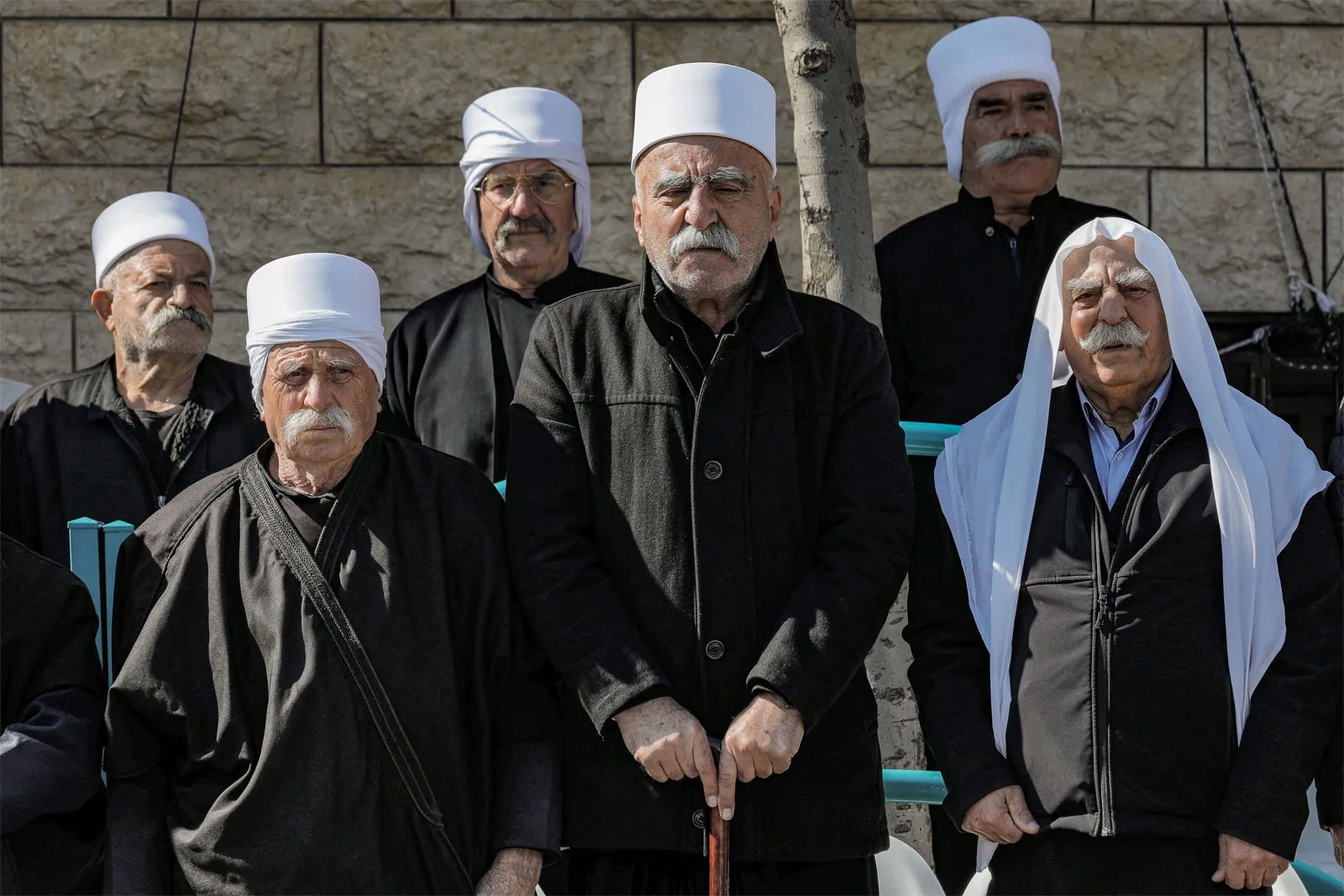 مجموعة من الرجال الدروز المسنين يرتدون الزي التقليدي الأسود والعمائم البيضاء يقفون أمام جدار حجري